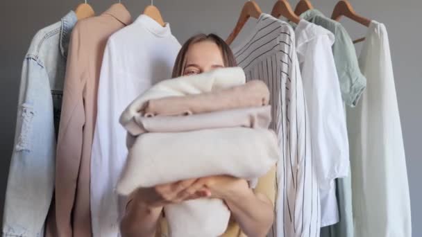 Shopping store. Discounted clothing. Clothing storage. Pretty smiling attractive young adult woman sore assistant posing near outfit rail offering stack of clothing from new collection. - Video, Çekim