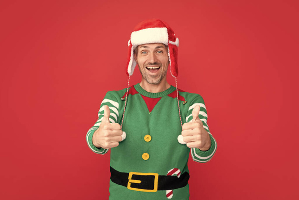 Elf kostümlü, hayret verici bir adam. Kırmızı arka planda Noel Baba şapkalı xmas adamı. Mutlu yıllar. Mutlu noeller. başparmak yukarı. - Fotoğraf, Görsel