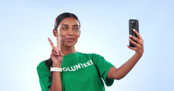 Mujer voluntaria, selfie y sonríe en el estudio con signo de paz, emoji y orgullo por el servicio comunitario con fondo azul. Chica, trabajador de ONG y feliz con el icono, revisión y v para ganar en las redes sociales. - Imágenes, Vídeo