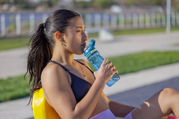 Λατινική κοπέλα σε αθλητικά ρούχα πόσιμο νερό σε μια υπαίθρια μηχανή άσκησης. - Φωτογραφία, εικόνα