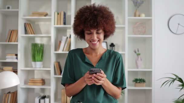 Akıllı telefon kullanan ve evde gülümseyen mutlu Afro-Amerikan kıvırcık kadın portresi.  - Video, Çekim