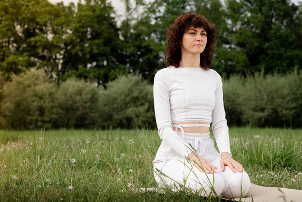 Concept of calmness. Sports woman sitting on yoga mat put hut on her lap. Female focused on breathing at the park. Full concentration mindfulness. Zen posture. High quality photo - Photo, Image
