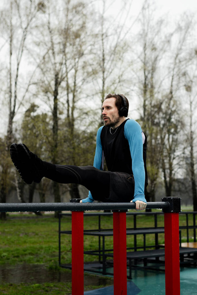 Sportsman balancing legs up using parallel bars. Powerful young man doing l sits abs over metal grips. Fitness training on sports court. Gymnastics exercise in park. High quality photo - Photo, Image
