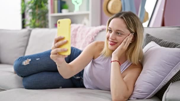 Νεαρή ξανθιά γυναίκα που παίρνει selfie φωτογραφία με smartphone που βρίσκεται στον καναπέ στο σπίτι - Πλάνα, βίντεο