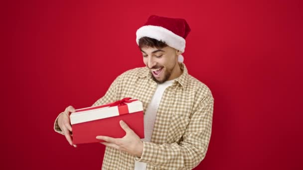 クリスマスの帽子を身に着けている若いアラブの男は,孤立した赤い背景の上に動揺しているように見えるギフトを荷を解く - 映像、動画