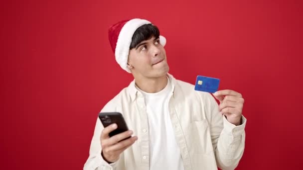 Jonge Spaanse man winkelen met smartphone en creditcard dragen kerstmuts over geïsoleerde rode achtergrond - Video