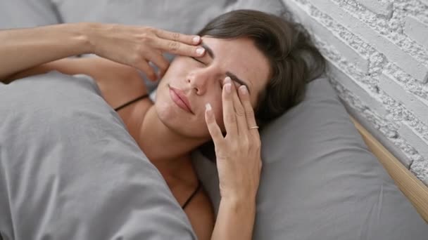 Mooie jonge Spaanse vrouw, gestresst en lijdt aan hoofdpijn, liggend in bed in een slaapkamer, weerspiegelt haar moeilijke levensstijl - Video