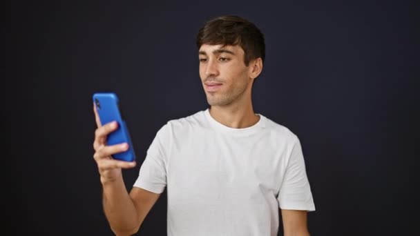 Χαρούμενος νεαρός Ισπανός με αυτοπεποίθηση χαμογελώντας ενώ στέκεται, μιλώντας στο τηλέφωνο και διασκεδάζοντας κατά τη διάρκεια μιας βιντεοκλήσης σε ένα δροσερό απομονωμένο φόντο μαύρου τοίχου - Πλάνα, βίντεο