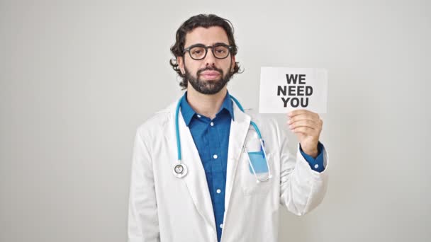 Young hispanic man doctor standing with serious expression holding we need you paper over isolated white background - Footage, Video