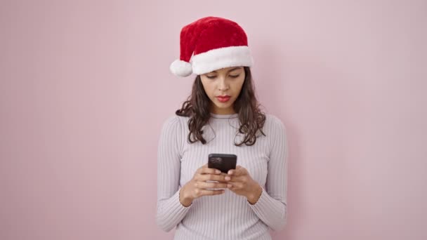 Jovem bela mulher hispânica usando chapéu de Natal usando smartphone sobre fundo rosa isolado - Filmagem, Vídeo