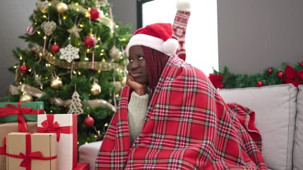 Afrykanka kobieta z plecionymi włosami siedzi na kanapie przez choinkę patrząc zdenerwowany w domu - Materiał filmowy, wideo