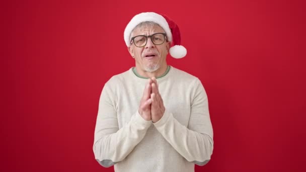 Μεσήλικας γκριζομάλλης που φοράει χριστουγεννιάτικο καπέλο και ζητάει τύχη σε απομονωμένο κόκκινο φόντο - Πλάνα, βίντεο