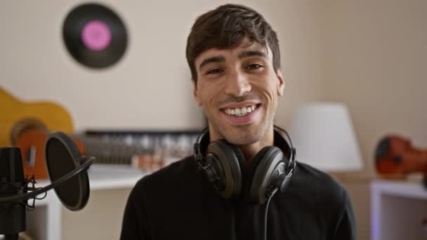 Joven y sonriente hispano irradia confianza mientras disfruta de su hobby musical en el estudio, con los auriculares puestos, inmerso en sus melodías. - Metraje, vídeo