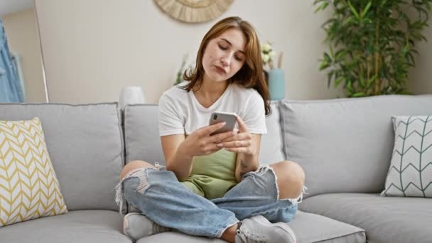 Νεαρή γυναίκα που χρησιμοποιεί το smartphone κάθεται στον καναπέ αναζητούν αναστατωμένος στο σπίτι - Πλάνα, βίντεο