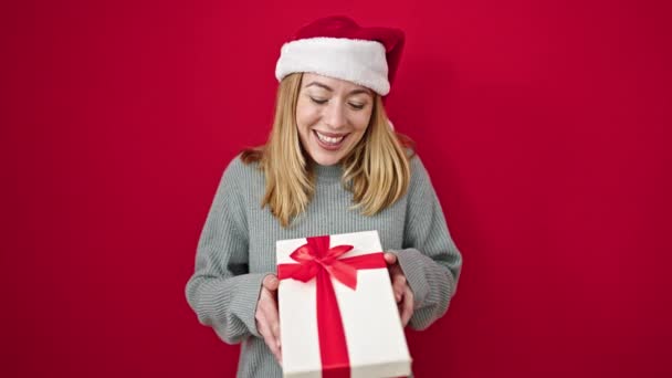 Jeune femme blonde souriante confiante tenant cadeau de Noël sur fond rouge isolé - Séquence, vidéo