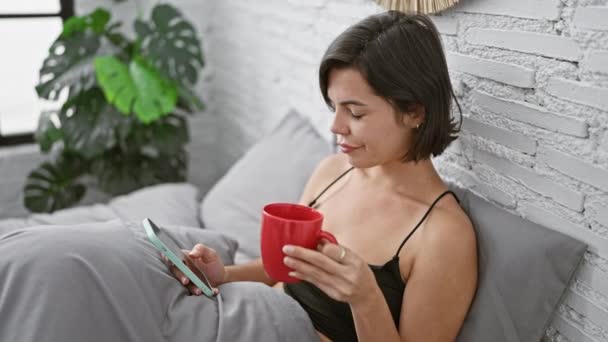 Poranne wibracje, szczęśliwa młoda Latynoska kobieta wygodnie siedząca na łóżku w sypialni, pijąca kawę, pijąca wiadomość na swoim smartfonie z pewnym uśmiechem. - Materiał filmowy, wideo