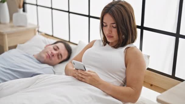 Beautiful couple lying on bed using smartphone while boyfriend sleep at bedroom - Footage, Video