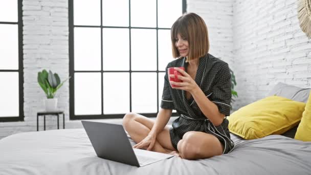 Молода красива іспаномовна жінка робить відеодзвінок з ноутбуком, що сидить на ліжку п'є каву в спальні - Кадри, відео