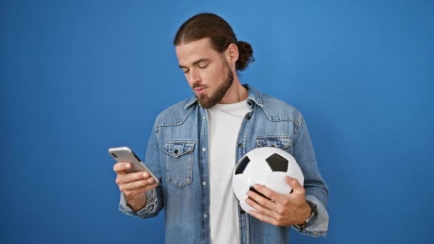Молодий іспаномовний чоловік тримає футбольний м'яч і смартфон, який виглядає засмученим на ізольованому синьому фоні - Кадри, відео