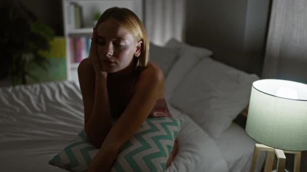 Mujer rubia joven sentada en la cama sufriendo por el dolor cervical en el dormitorio - Imágenes, Vídeo