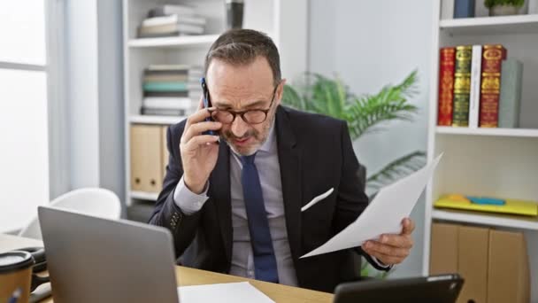 Energický muž středního věku s šedivými vlasy, ponořený do své profesionální role v kanceláři, sebevědomě mluví na smartphonu, zatímco pozorně čte zásadní obchodní dokument. - Záběry, video