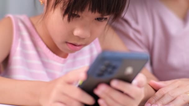 Onnellinen aasialainen perhe äiti ja söpö tytär käyttävät puhelinta yhdessä kotona. Äiti ja pieni tyttö nauraa iloisesti älypuhelimella katsomassa hauska sosiaalisen median video app istuu pöydässä yhdessä. - Materiaali, video