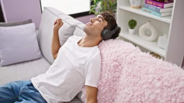 Retrato de un joven hispano relajado y seguro de sí mismo, disfrutando alegremente del sonido de la música, escuchando a través de auriculares, sentado en el interior del sofá en un acogedor y moderno apartamento. - Imágenes, Vídeo