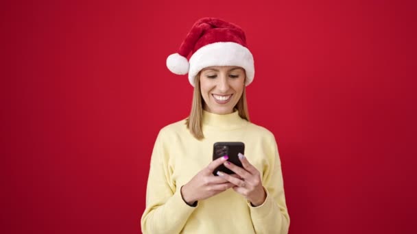 Giovane donna bionda che indossa il cappello di Natale utilizzando smartphone su sfondo rosso isolato - Filmati, video