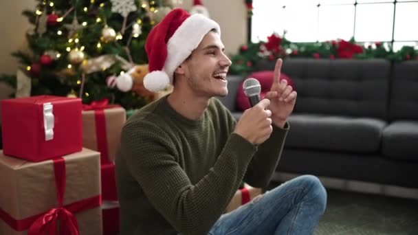 Giovane uomo ispanico cantare canzone seduta sul pavimento da albero di Natale a casa - Filmati, video