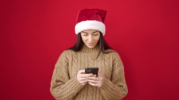 Jovem bela mulher hispânica vestindo chapéu de Natal apontando smartphone tela com expressão surpresa sobre fundo vermelho isolado - Filmagem, Vídeo