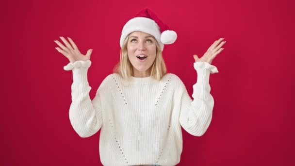 Mujer rubia joven expresión sorpresa usando sombrero de Navidad sobre fondo rojo aislado - Imágenes, Vídeo