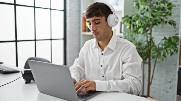 Magabiztos fiatal spanyol férfi élvezi a zenét, miközben ügyesen dolgozik laptop az irodában - Felvétel, videó
