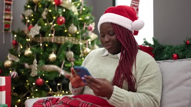 Mulher africana com cabelo trançado usando smartphone sentado na árvore de natal em casa - Filmagem, Vídeo