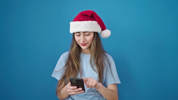 Giovane bella donna ispanica indossa cappello di Natale che punta a smartphone con espressione vincitore su isolato sfondo blu - Filmati, video