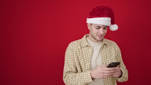 Fiatal spanyol férfi visel karácsonyi kalap okostelefon néz oldalra, mint az elszigetelt piros háttér - Felvétel, videó