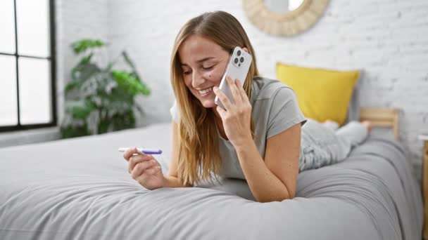 Jeune femme blonde dans une conversation heureuse sur son téléphone, couchée détendue au lit avec un test de grossesse tôt le matin un nouveau chapitre lumineux dans sa chambre confortable. - Séquence, vidéo
