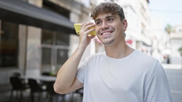 Молодий іспаномовний чоловік слухає голосове повідомлення, посміхаючись на терасі кав'ярні - Кадри, відео