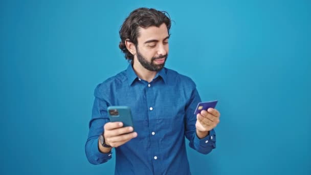 Junger hispanischer Mann mit Smartphone und Kreditkarte vor isoliertem blauen Hintergrund - Filmmaterial, Video