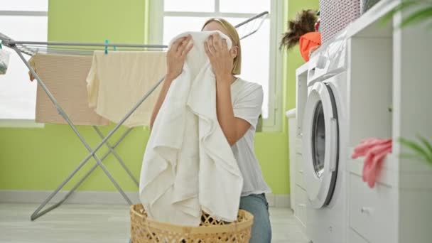 Mujer rubia joven oliendo toalla limpia sonriendo en la lavandería - Imágenes, Vídeo