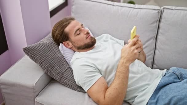 Jonge Spaanse man met behulp van smartphone liggend op de bank thuis - Video