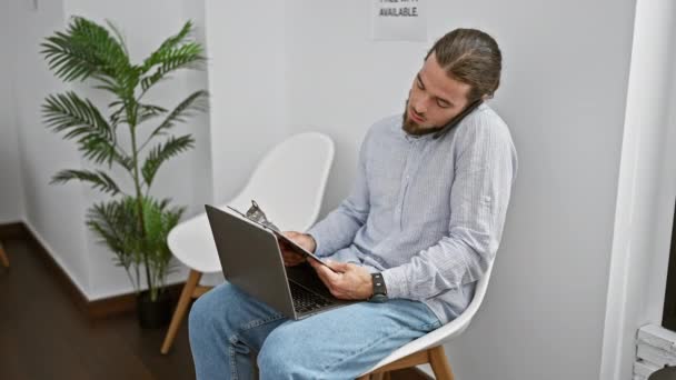 Молодий іспаномовний чоловік розмовляє на смартфоні читаючи документ, сидячи на стільці в залі очікування - Кадри, відео