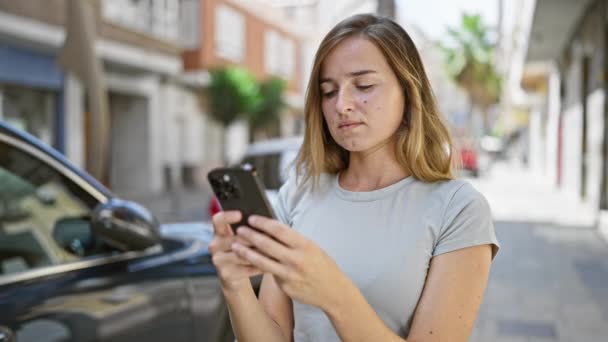 Весела молода блондинка щасливо торкається свого смартфона, веде цифрову розмову на сонячній міській вулиці - Кадри, відео