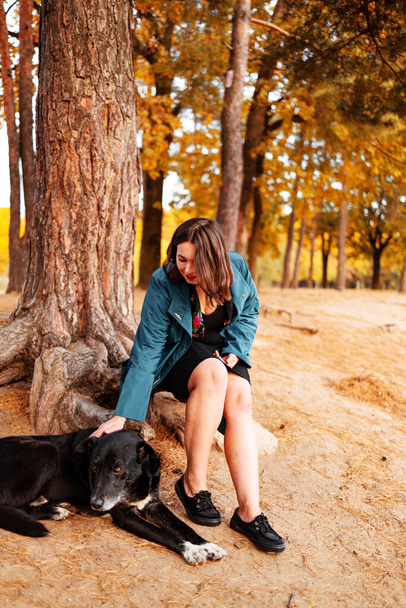 Lány és a kisállat túrázás és egy kis pihenés a tó közelében. Kék kabátos lány a nagy fekete kutyájával az őszi erdőben ül a fatörzsön.. - Fotó, kép