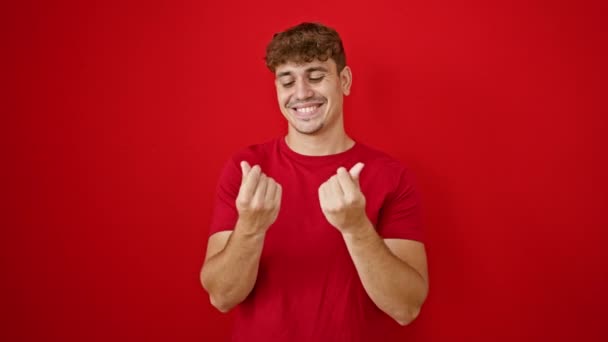 Un joven hispano confiado hace alegremente un gesto de "gastar dinero", parado sobre un fondo rojo aislado, sus manos expresando alegría, diversión y confianza, su sonrisa gritando felicidad - Metraje, vídeo