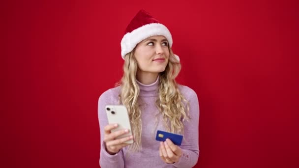 Junge blonde Frau kauft mit Smartphone und Kreditkarte ein und trägt Weihnachtsmütze über rotem Hintergrund - Filmmaterial, Video