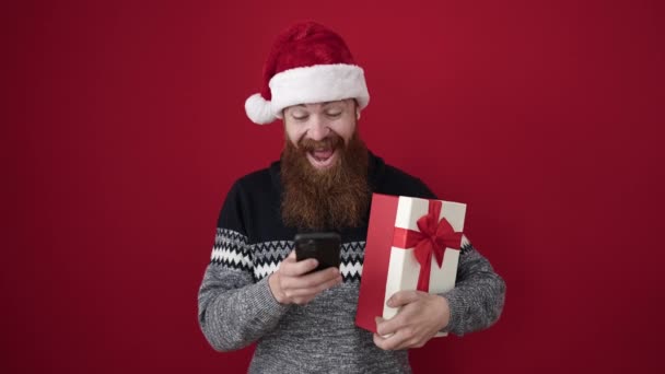 Giovane rossa uomo in possesso di regalo di Natale utilizzando smartphone su sfondo rosso isolato - Filmati, video