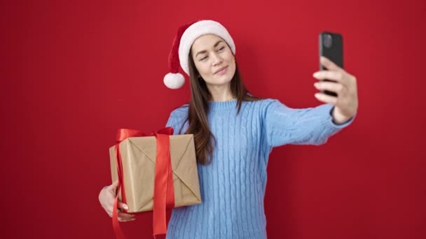 Nuori valkoihoinen nainen yllään joulu hattu tehdä selfie älypuhelimella tilalla lahja yli eristetty punainen tausta - Materiaali, video