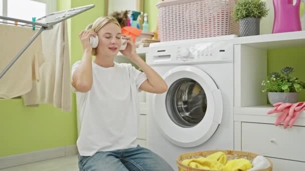 Молода блондинка слухає музику, тримає одяг кошика в пральні - Кадри, відео