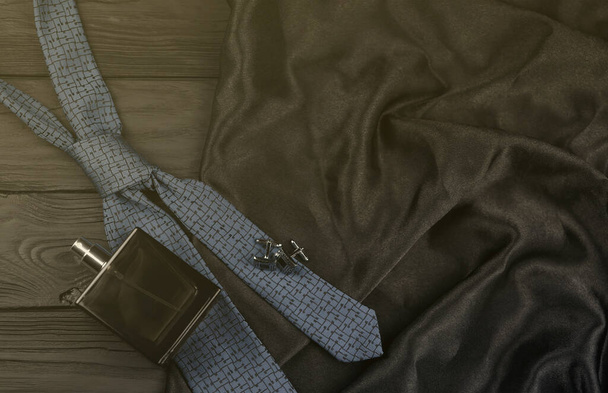 Uma garrafa de colônia mens e abotoaduras com gravata azul jaz em um fundo de tecido de luxo preto em uma mesa de madeira. Mens acessórios clássicos. DOF superficial - Foto, Imagem