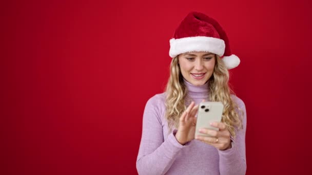 fiatal szőke nő visel karácsonyi kalap segítségével smartphone meglepő kifejezés izolált piros háttér - Felvétel, videó